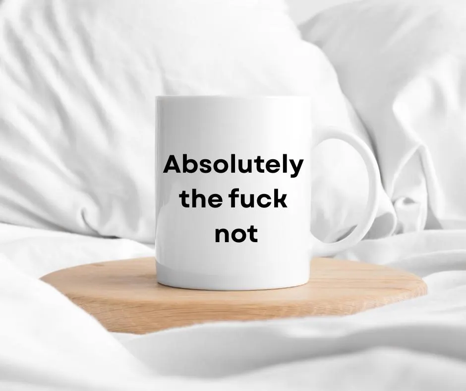 White mug with text 'Absolutely the fuck not' on a wooden coaster in bed with white fabric background