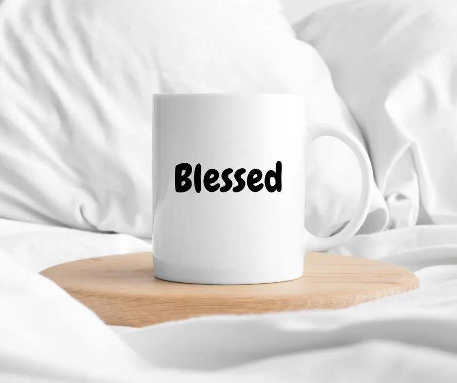 White mug with 'Blessed' text on a wooden coaster in bed with white fabric background