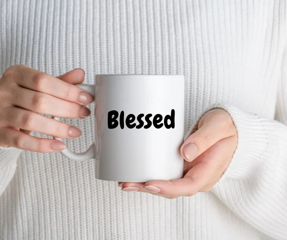 White mug with 'Blessed' text held by hands against a white sweater background