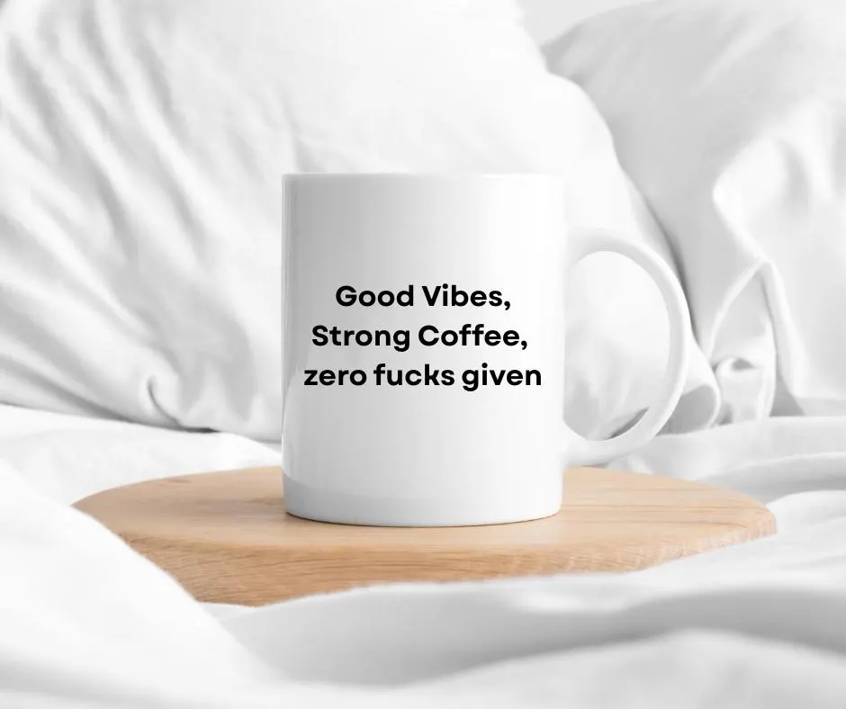 White mug with text that says "Good Vibes, Strong Coffee, zero fucks given" on a wooden coaster in bed with white fabric background