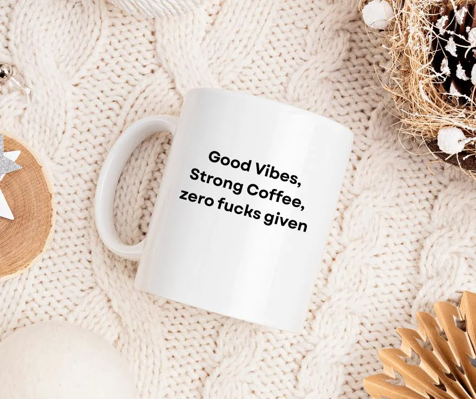 White mug with text 'Good Vibes, Strong Coffee, zero fucks given' on a textured surface.