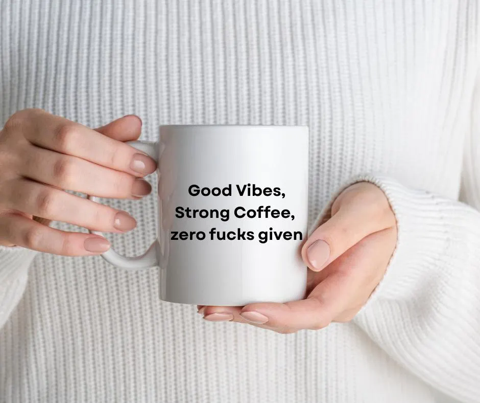 Person holding a white mug with text 'Good Vibes, Strong Coffee, zero fucks given' against a white sweater background