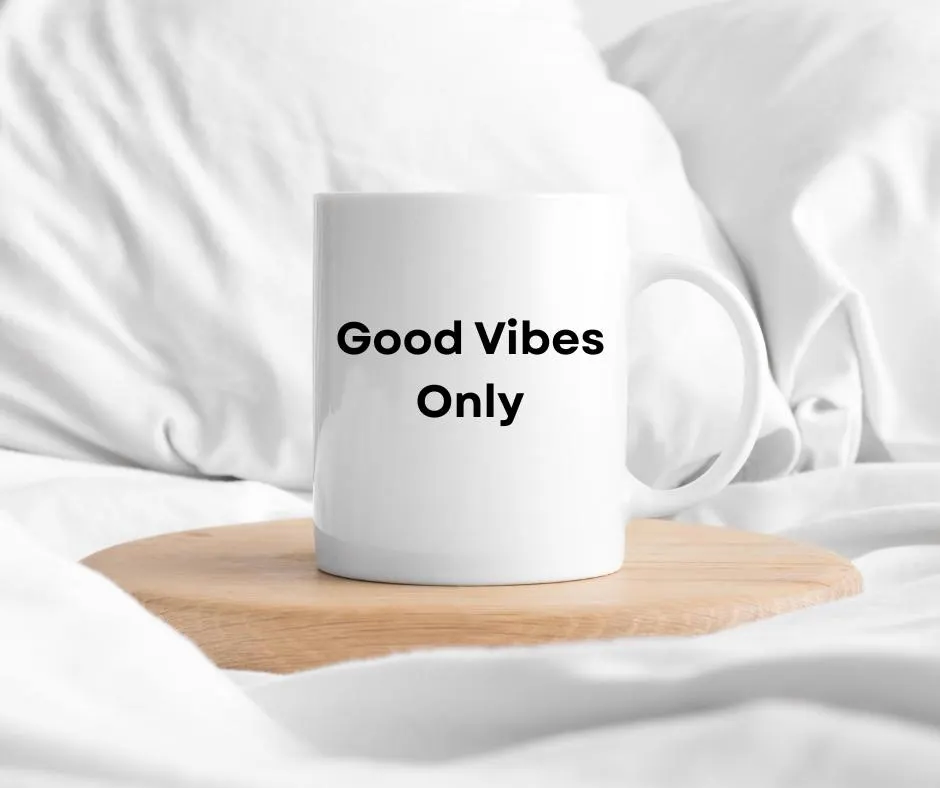 White mug with 'Good Vibes Only' text on a wooden coaster in bed with white fabric background