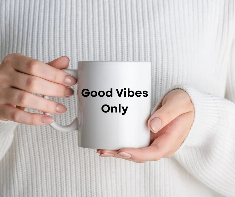 White mug with 'Good Vibes Only' text held by hands against a white sweater background
