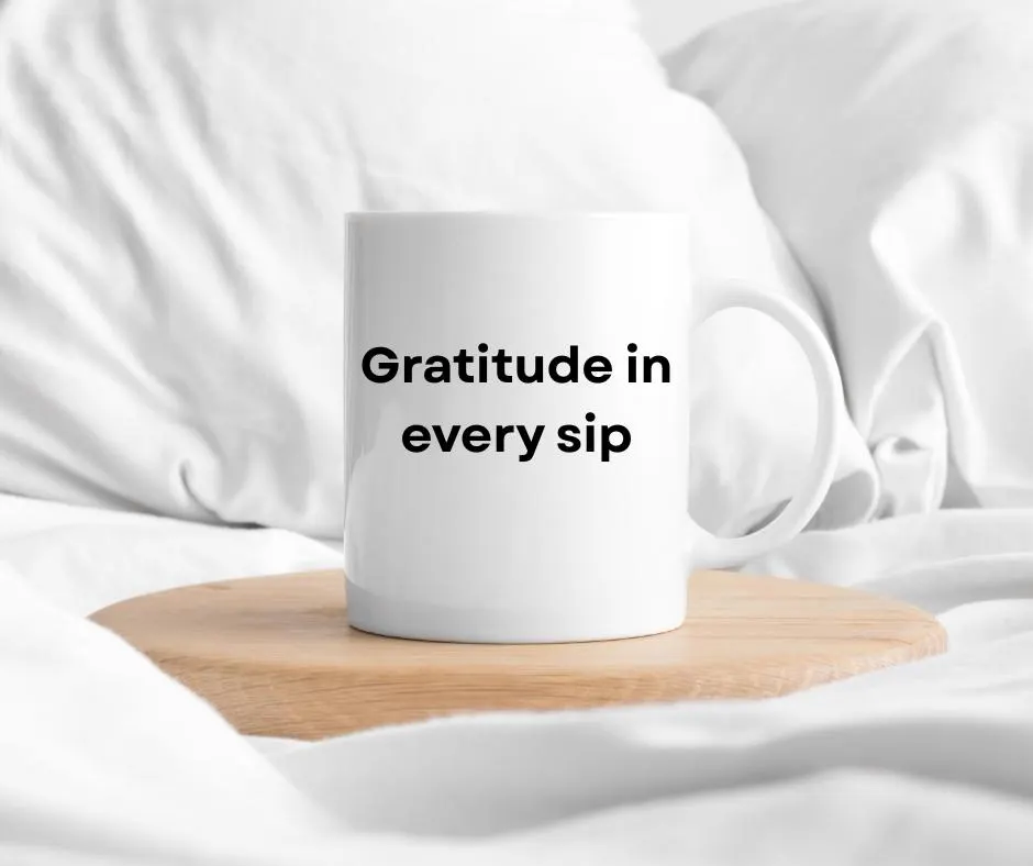 White mug with 'Gratitude in every sip' text on a wooden coaster in bed with white fabric background