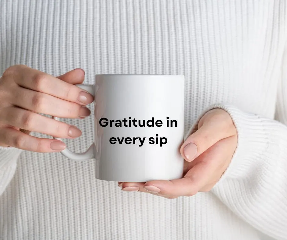 White mug with 'Gratitude in every sip' text held by hands against a white sweater background