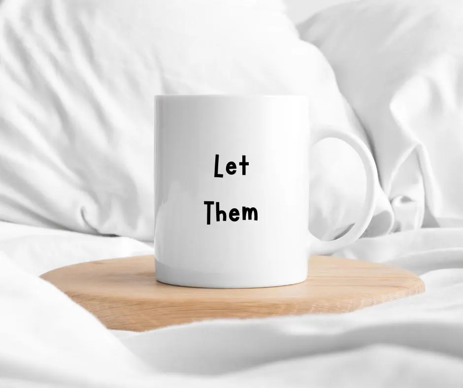 White mug with 'Let Them' text on a wooden coaster in bed with white fabric background