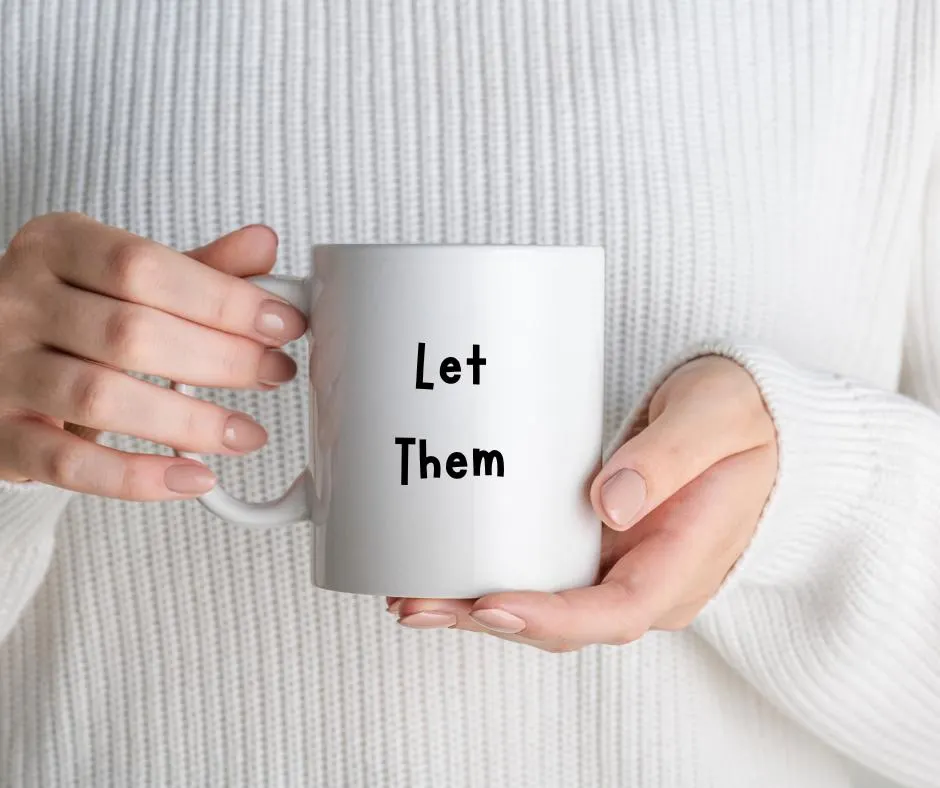 White mug with 'Let Them' text held by hands against a white sweater background