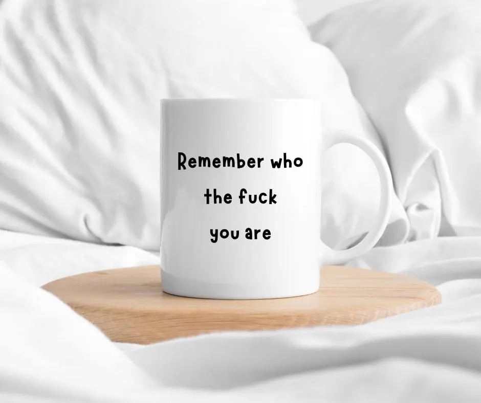 White mug with text 'Remember who the fuck you are' on a wooden coaster in a bed.