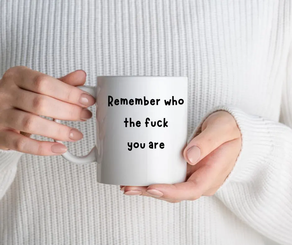 Person holding a white mug with text 'Remember who the fuck you are' against a white sweater background
