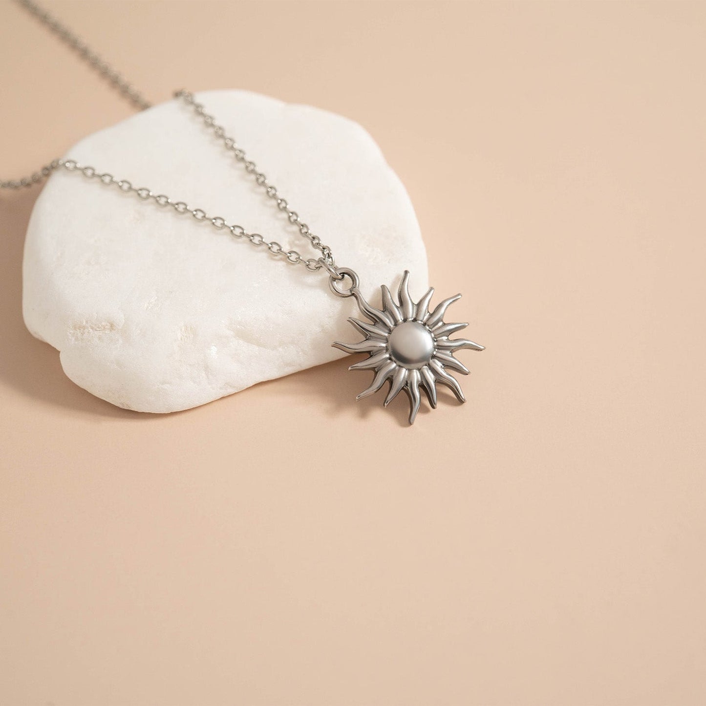 Silver sun-shaped pendant on a chain laying over a rock against a beige background