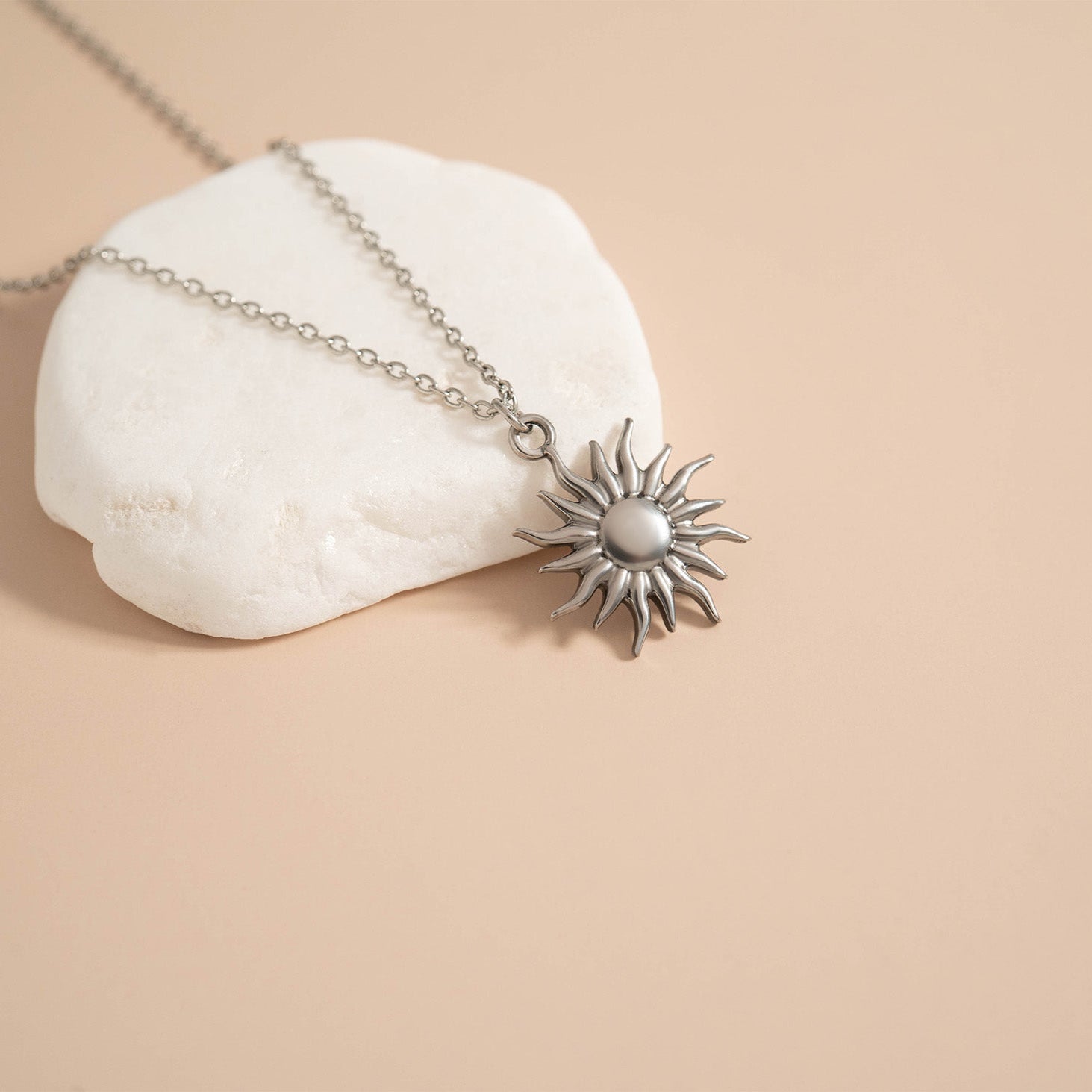 Silver sun-shaped pendant on a chain laying over a rock against a beige background