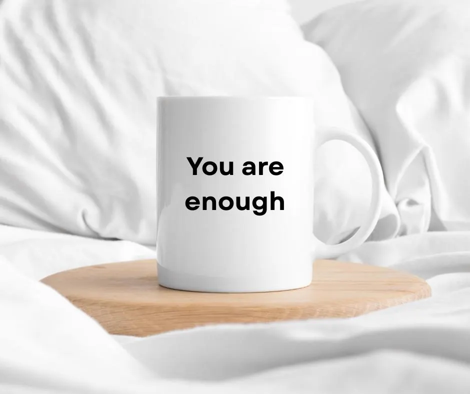 White mug with 'You are enough' text on a wooden coaster in bed with white fabric background