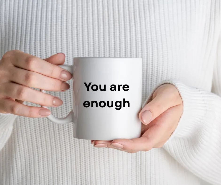 Person holding a white mug with 'You are enough' text against a white sweater background