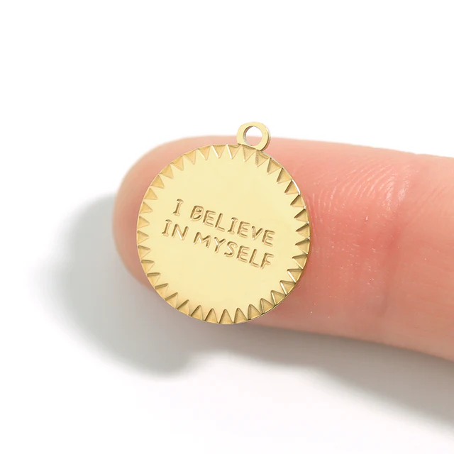 Gold pendant with 'I Believe In Myself' engraving held between fingers on a white background