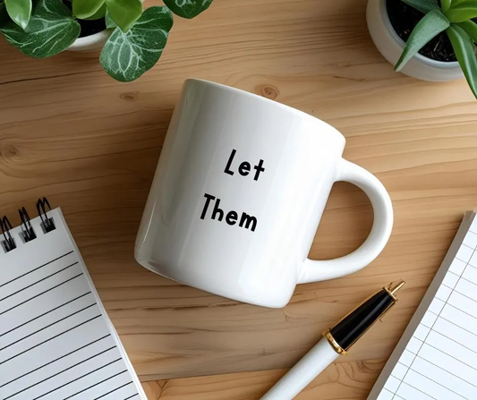 White mug with 'Let Them' text on a wooden desk with a notebook and pen.