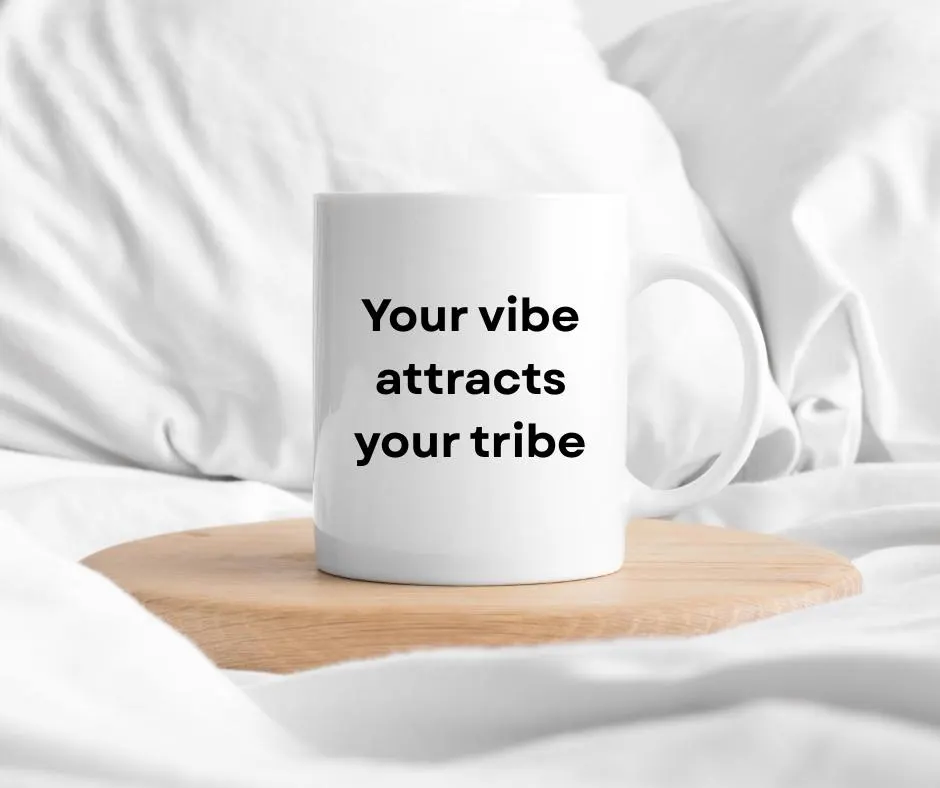 White mug with 'Your vibe attracts your tribe' text on a wooden coaster in bed with white fabric background