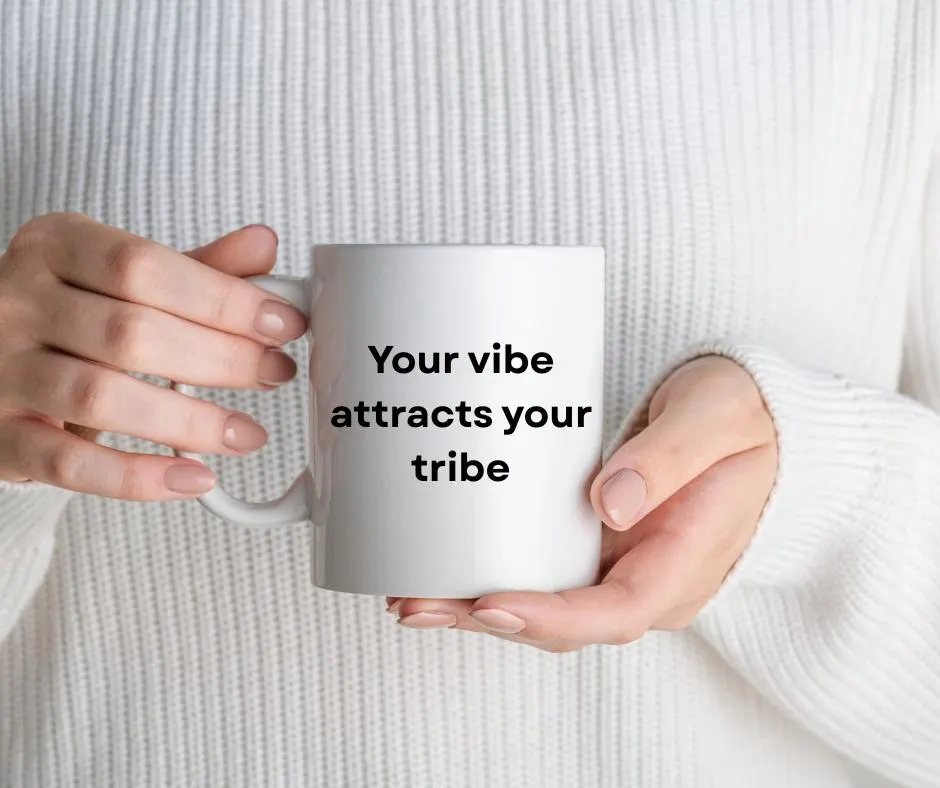 Person holding a white mug with text 'Your vibe attracts your tribe' against a white sweater background